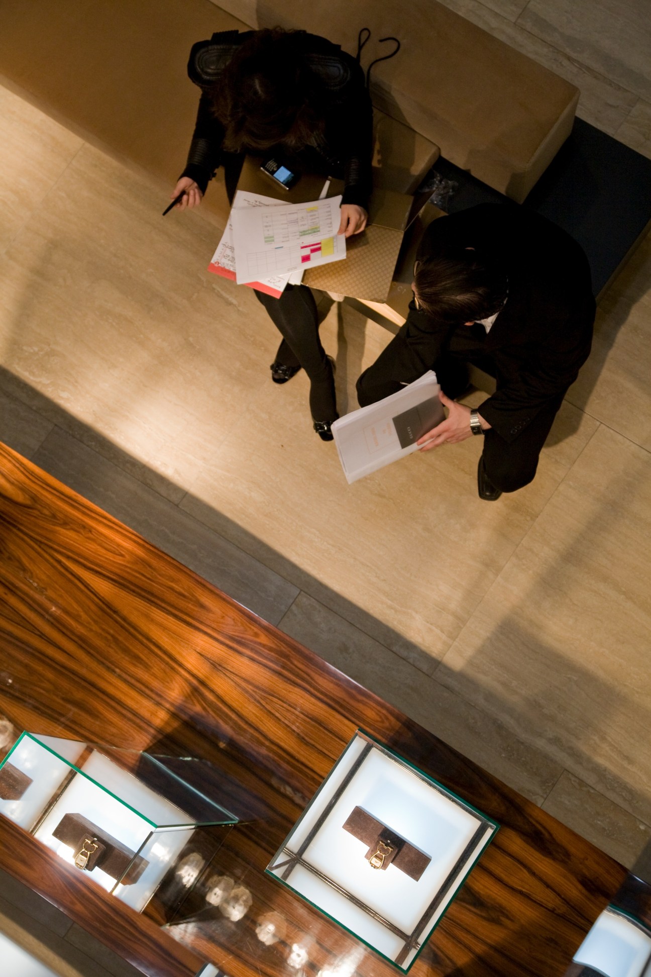 Overhead view of two people in dark business attire reviewing documents at a watch fair booth with display cases below.