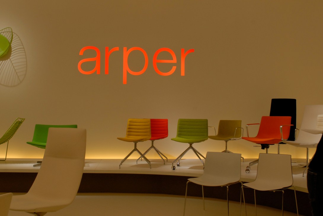 Arper showroom display featuring colorful modern chairs in green, yellow, red, and neutral tones with the Arper logo on the wall.