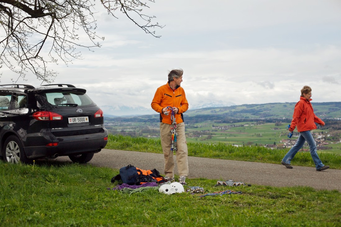 Bernhard Russi in orange jacket with climbing gear next to black Subaru Tribeca on hillside overlooking Swiss valley.