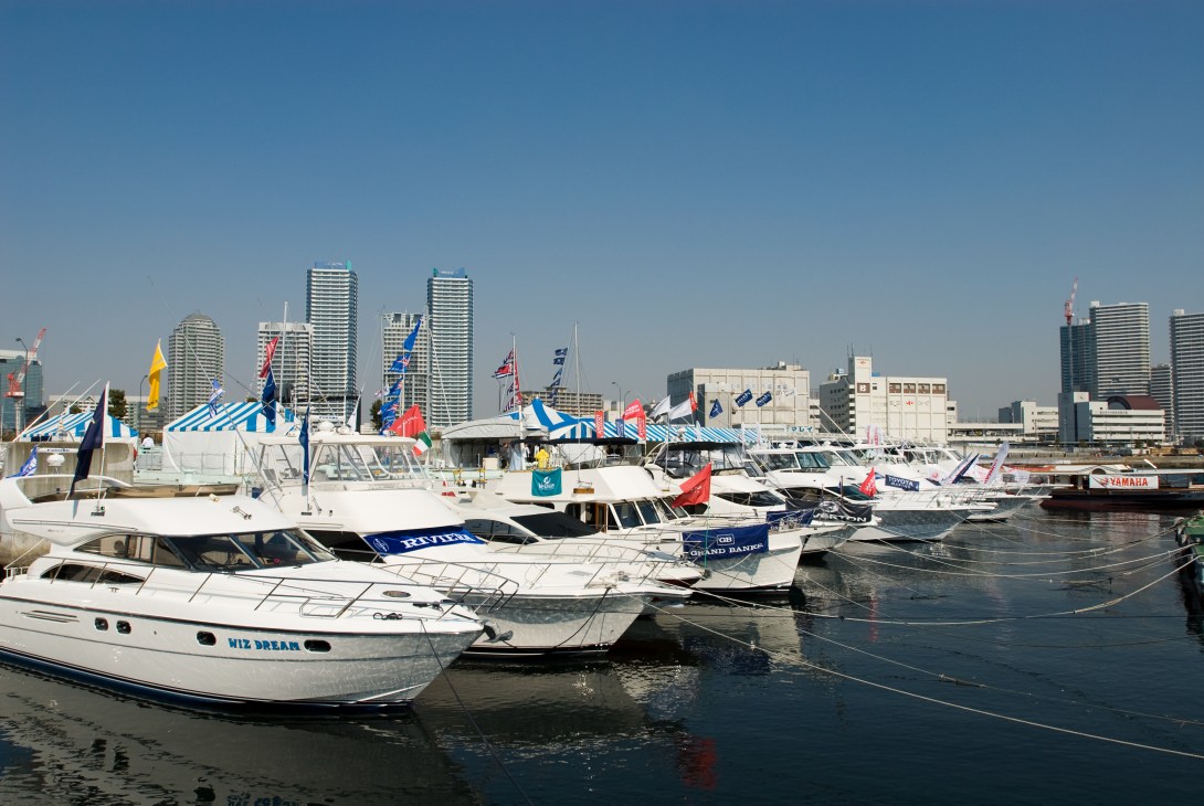 Multiple white and dark-hulled boats moored in Yokohama harbor with striped blue and white event tents and high-rise buildings in the background.