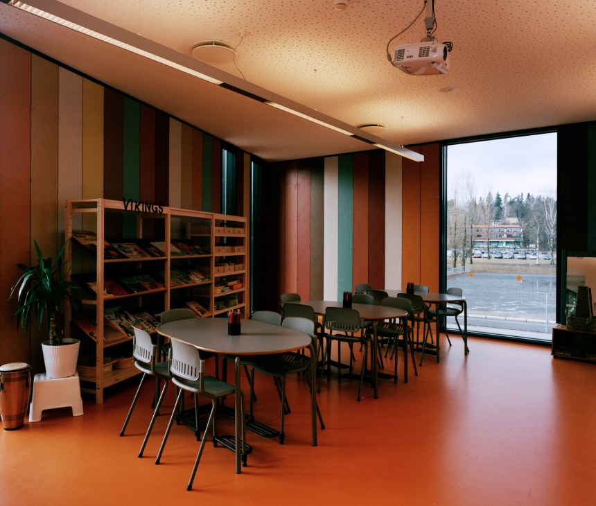 Colorful classroom with striped walls in red, green and tan, orange rubber flooring, wooden shelving and modern chairs around a table.
