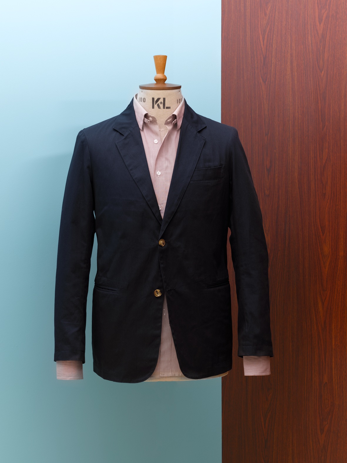 Navy blazer with pink shirt on tailoring mannequin against blue and wood-grain background.