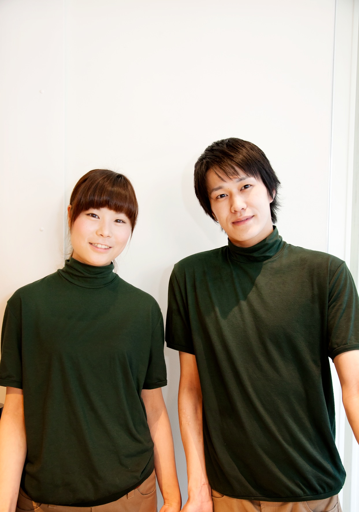 Two people wearing dark green turtleneck shirts pose against a white wall.