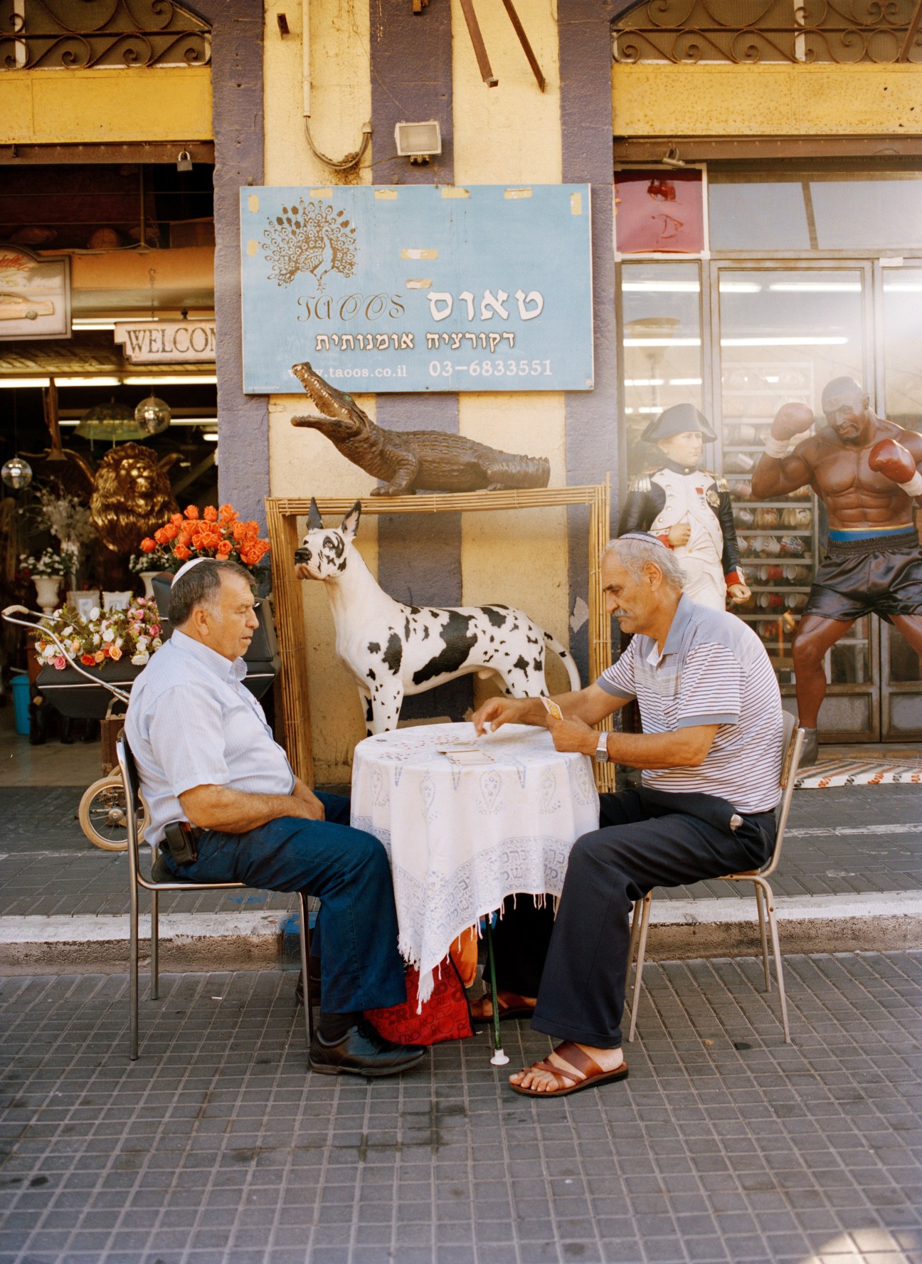 Two men play cards at a small table outside a shop in Jaffa's flea market, surrounded by eclectic decorations including animal sculptures and flowers.