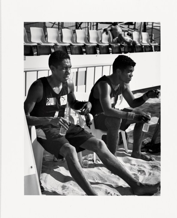 Two Timor Leste beach volleyball players sitting on sand in athletic wear, smiling and conversing.