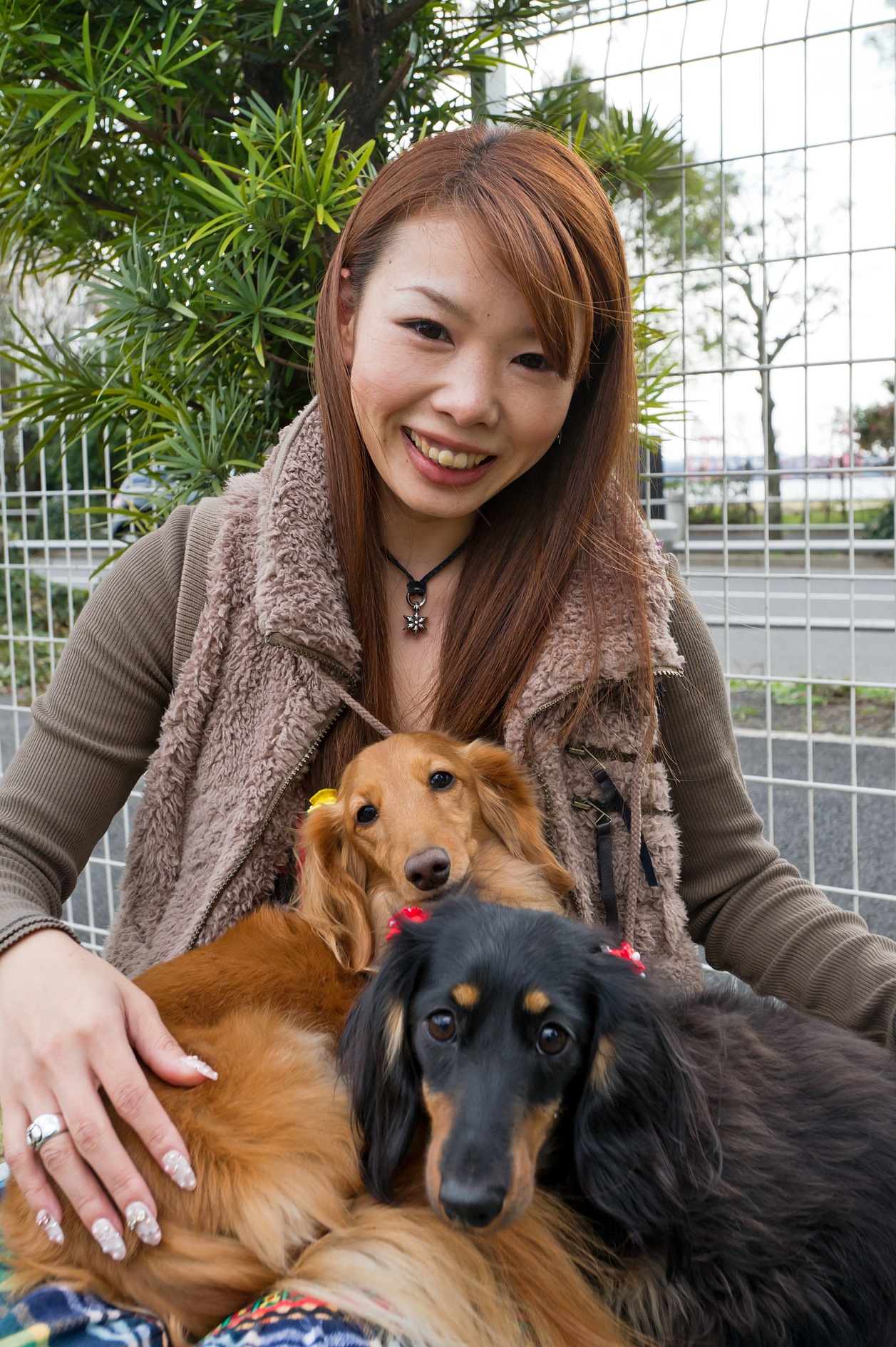 Woman smiling with two dogs, a golden-colored dog and a black and tan dog, outdoors with plants and glass panels behind them.