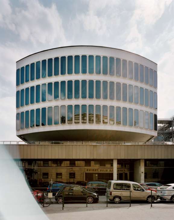 Cylindrical modernist building with curved white facade and blue-tinted windows, parked cars below.