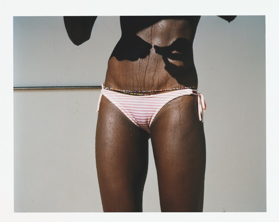 Shirtless male athlete with wet skin wearing pink and white striped swim briefs and beaded waist belt.