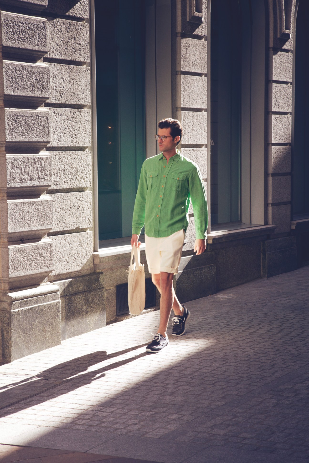 Man in green shirt and cream shorts walking through sunlit Lugano street with stone architecture.