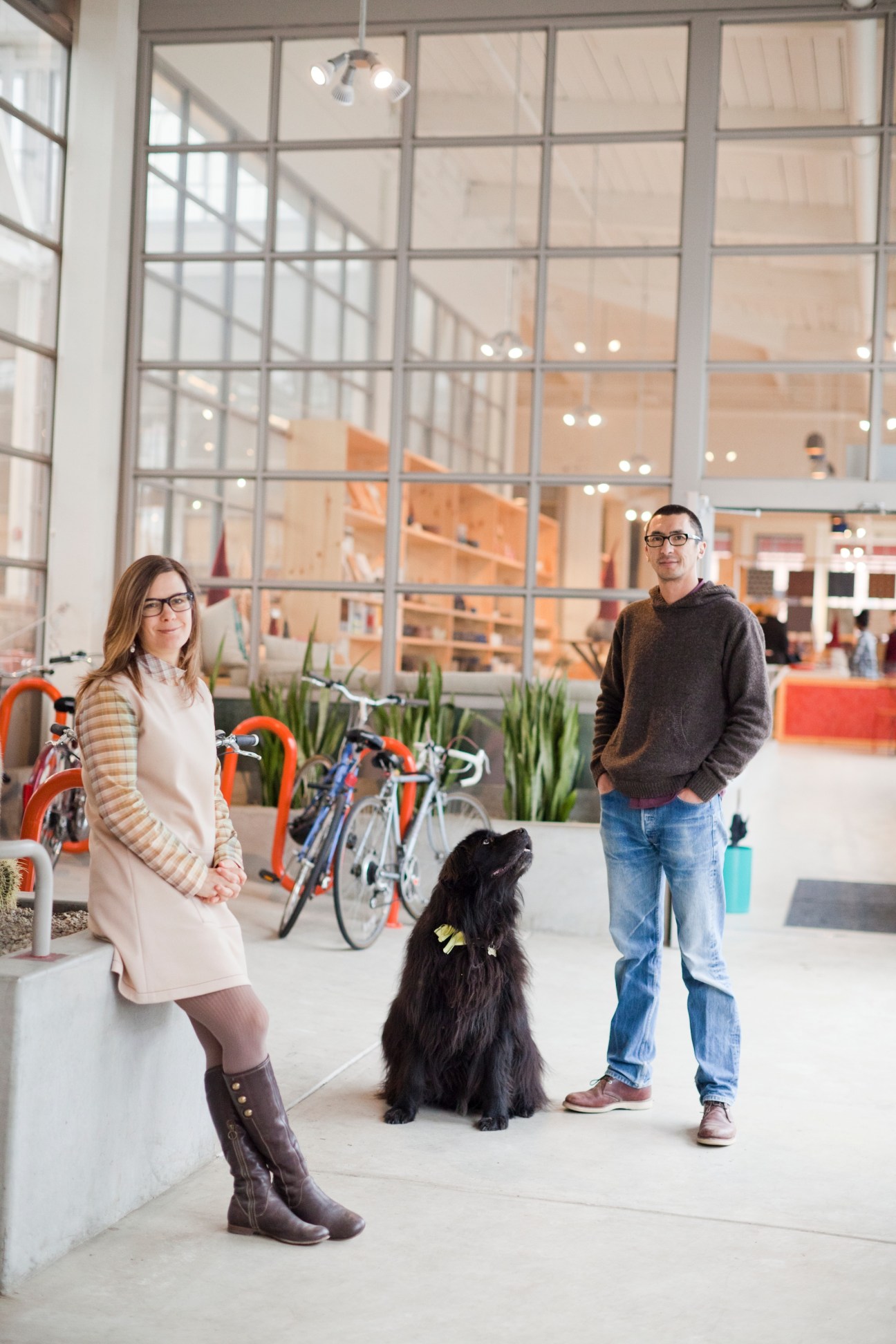 Robin Petravic and Catherine Bailey stand with a black dog in a bright industrial space with large windows and bicycles.