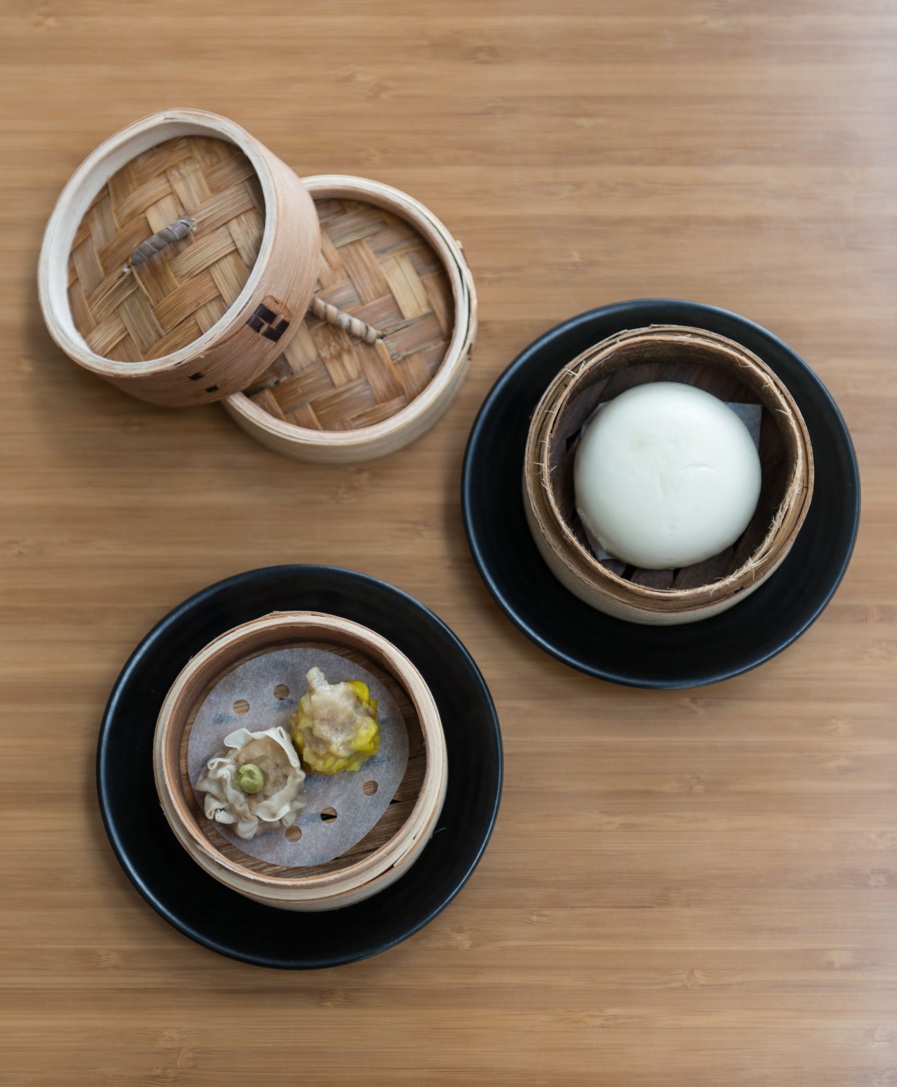Overhead view of bamboo steamer baskets with dim sum dumplings and buns on black plates against wooden surface