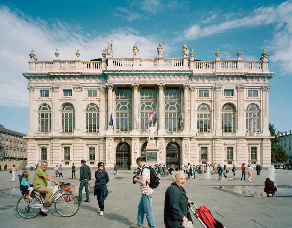 Palazzo Madama, a grand cream-colored Baroque palace with statues and columns, stands in a sunny plaza with pedestrians and cyclists.