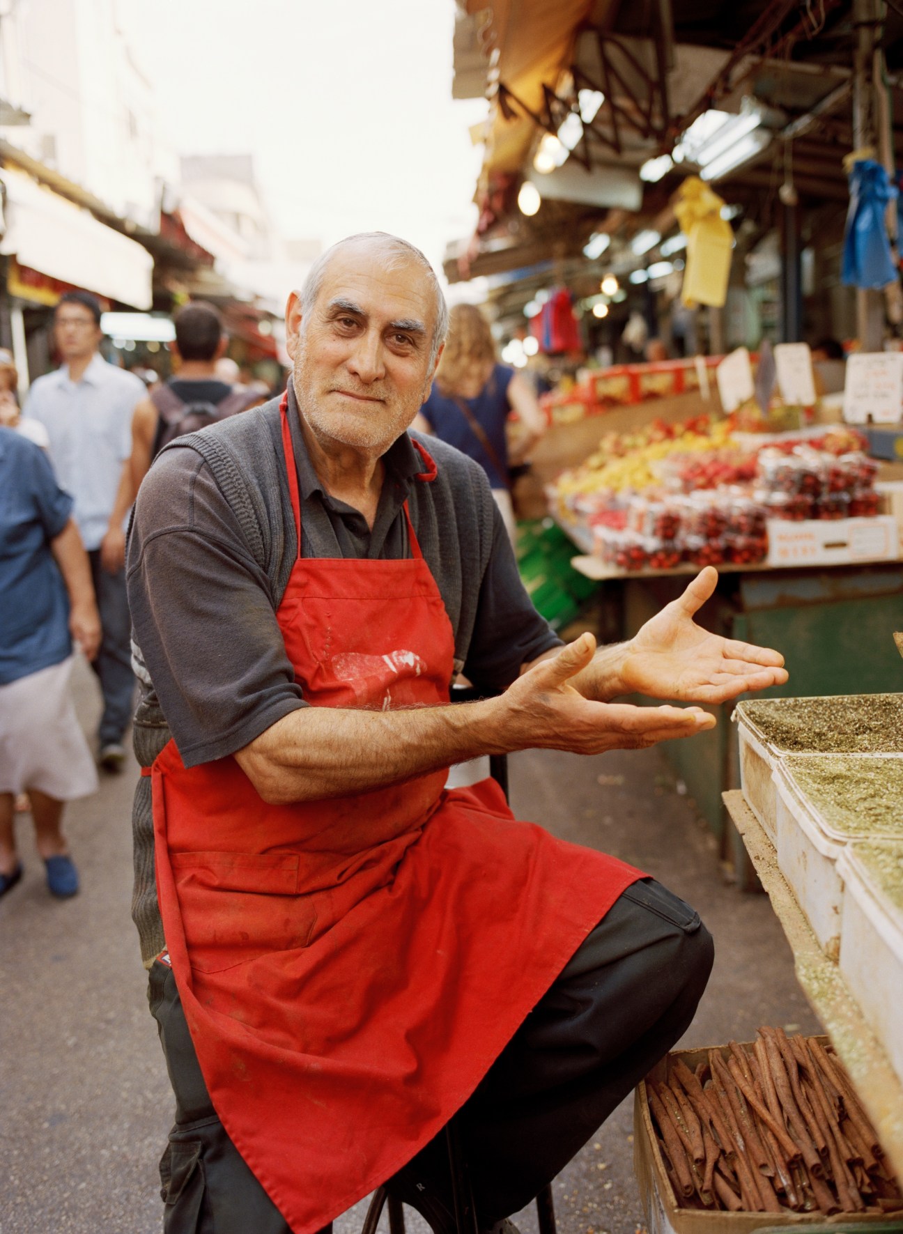 Elderly man in red apron gestures welcomingly at Carmel food market stall.