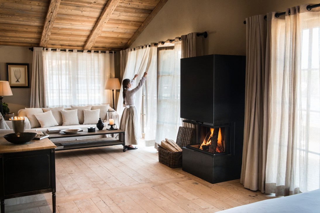 Luxury chalet interior at San Luis with fireplace, wooden beams, cream furnishings and woman adjusting curtains.