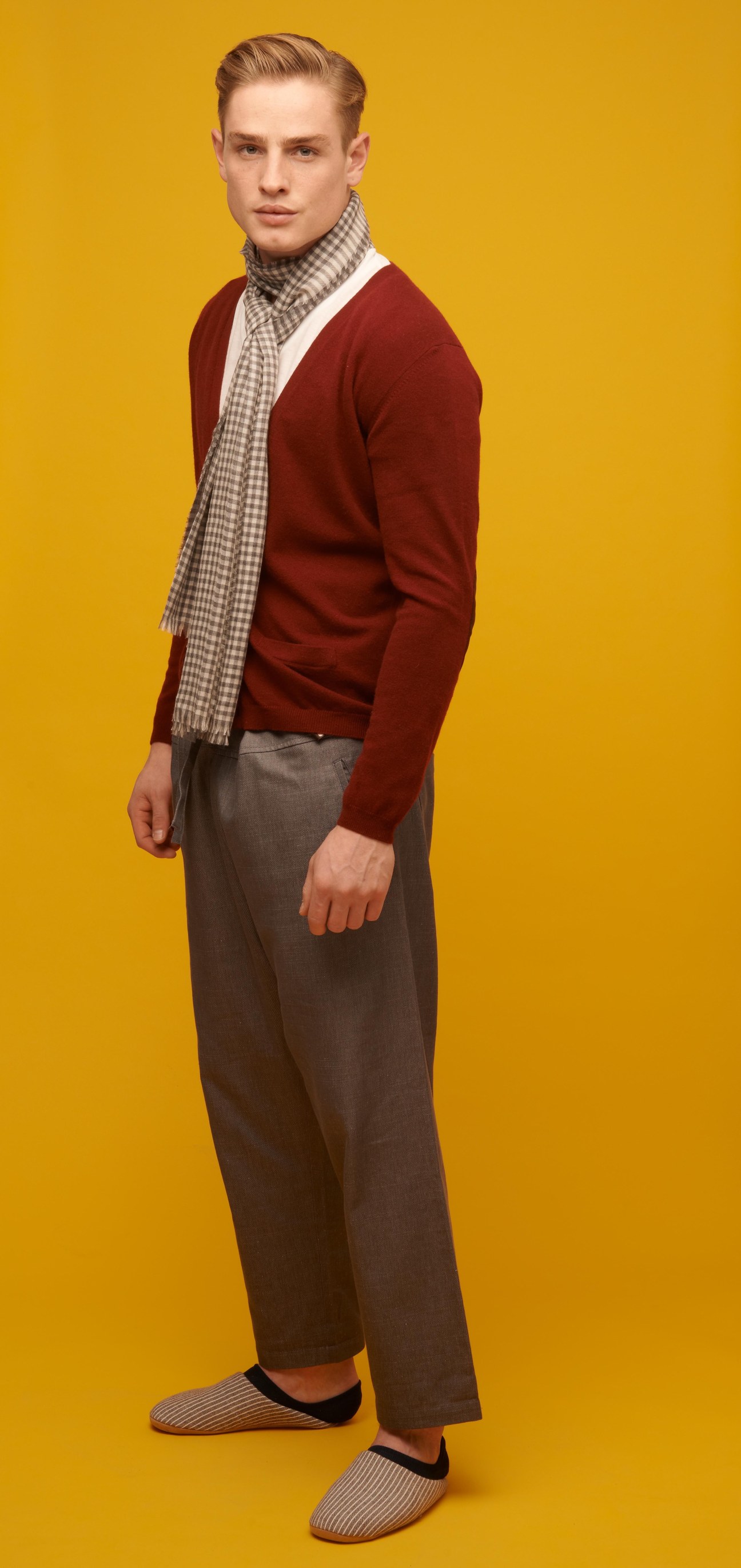 Man wearing burgundy cardigan, checkered scarf, gray trousers, striped socks and slippers against yellow background.