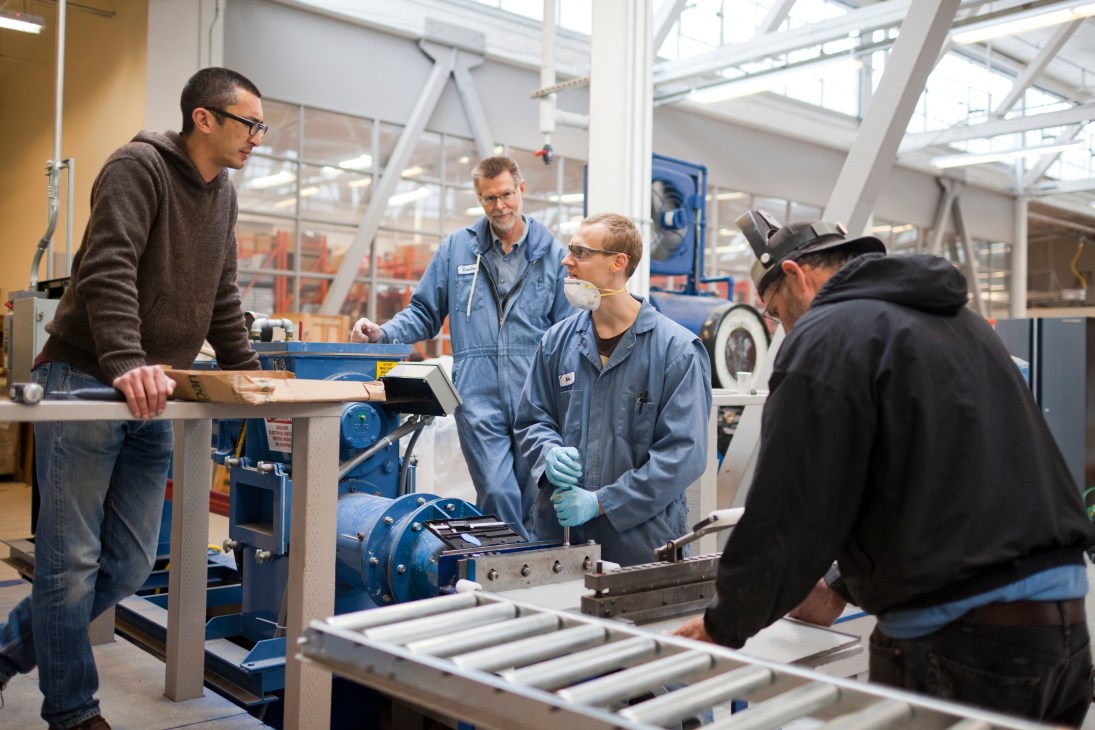 Industrial manufacturing workshop with workers gathered around machinery and equipment in a bright, naturally-lit space.