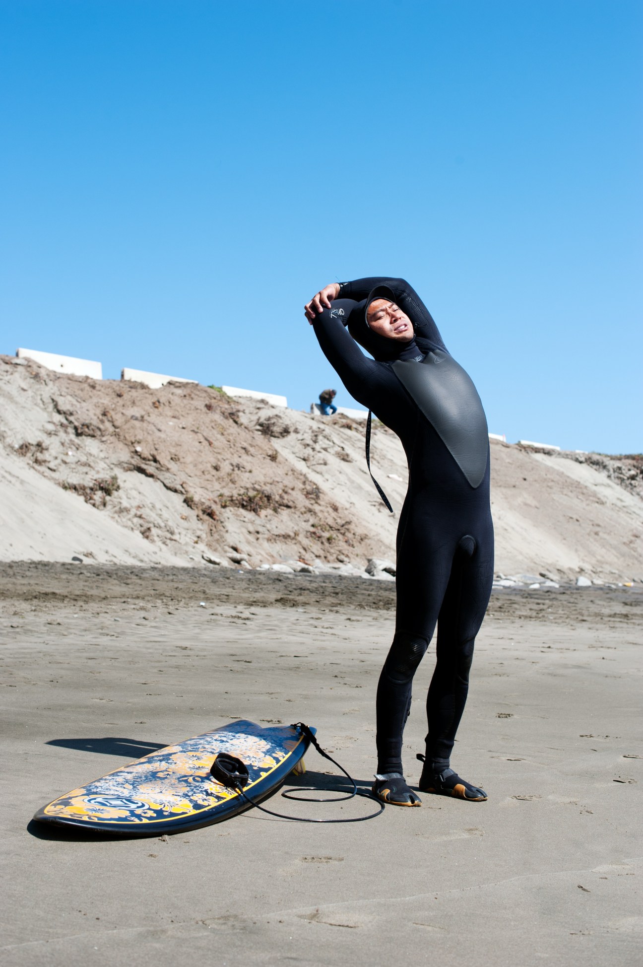 Surfer in black wetsuit adjusting hood on San Francisco beach with surfboard.