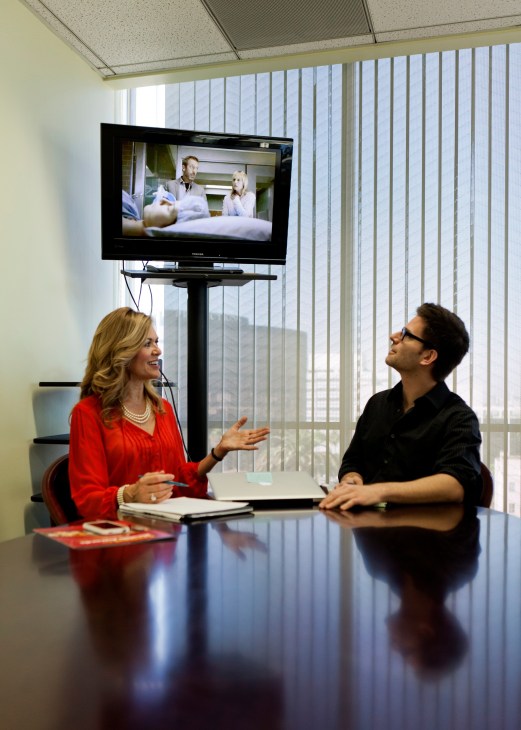 Woman in red shirt and man in black shirt review a medical scene on a TV monitor in an office