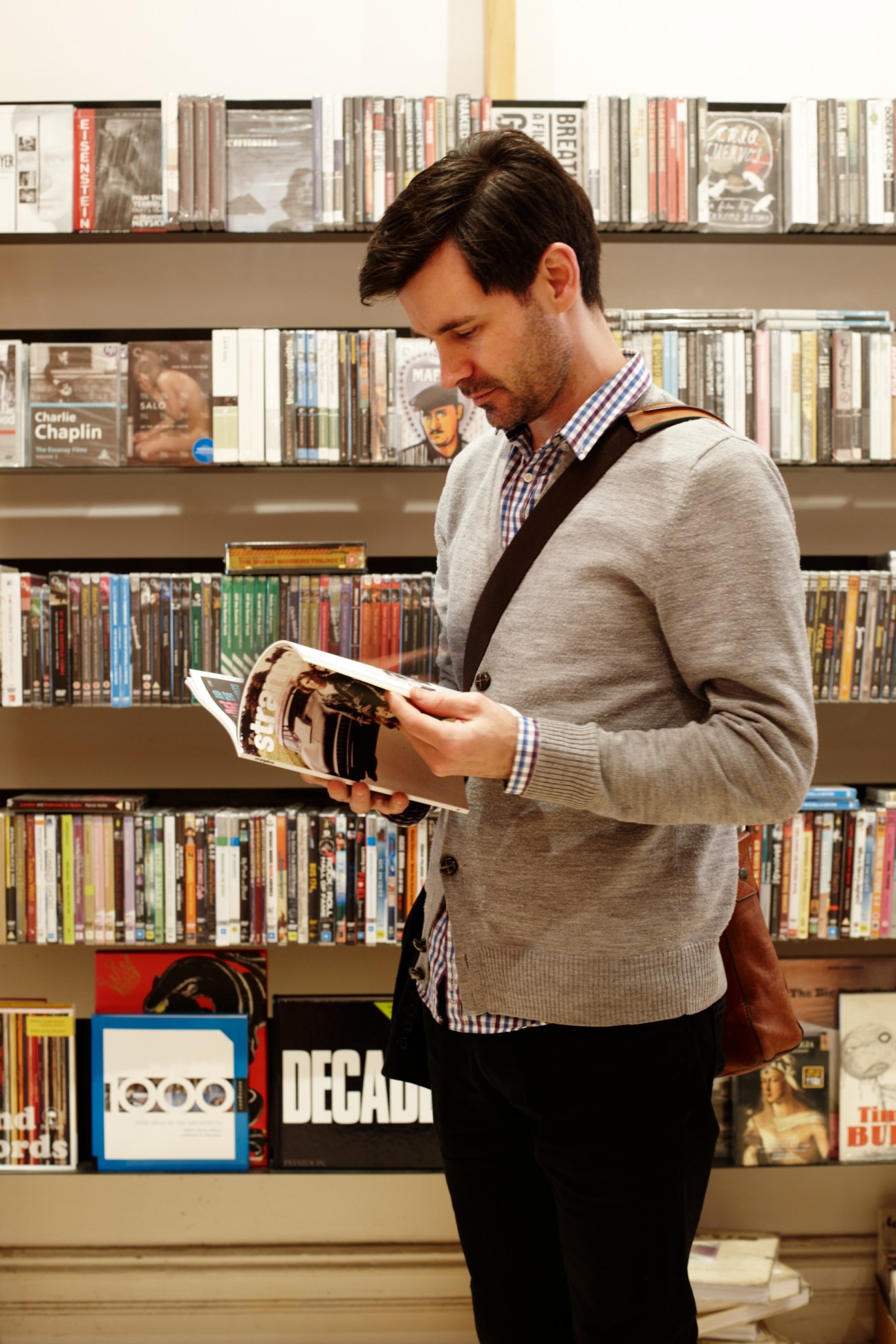 Man browsing a magazine at TITLE store with shelves of CDs and DVDs behind him.