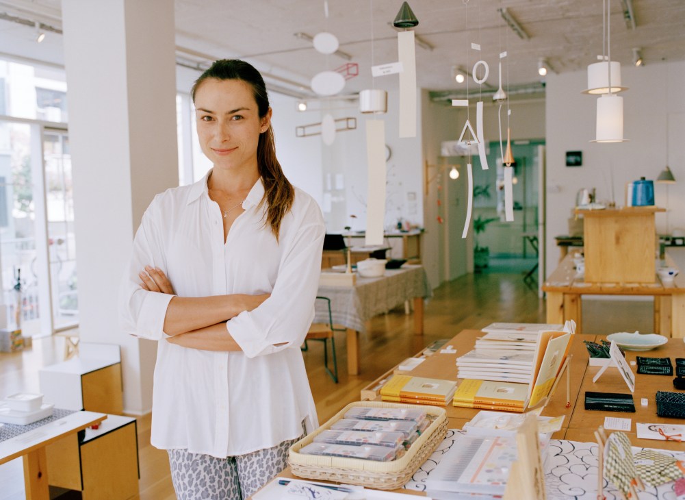 Woman in white shirt standing with arms crossed in a bright, modern office space with wooden furniture and design materials on desk.