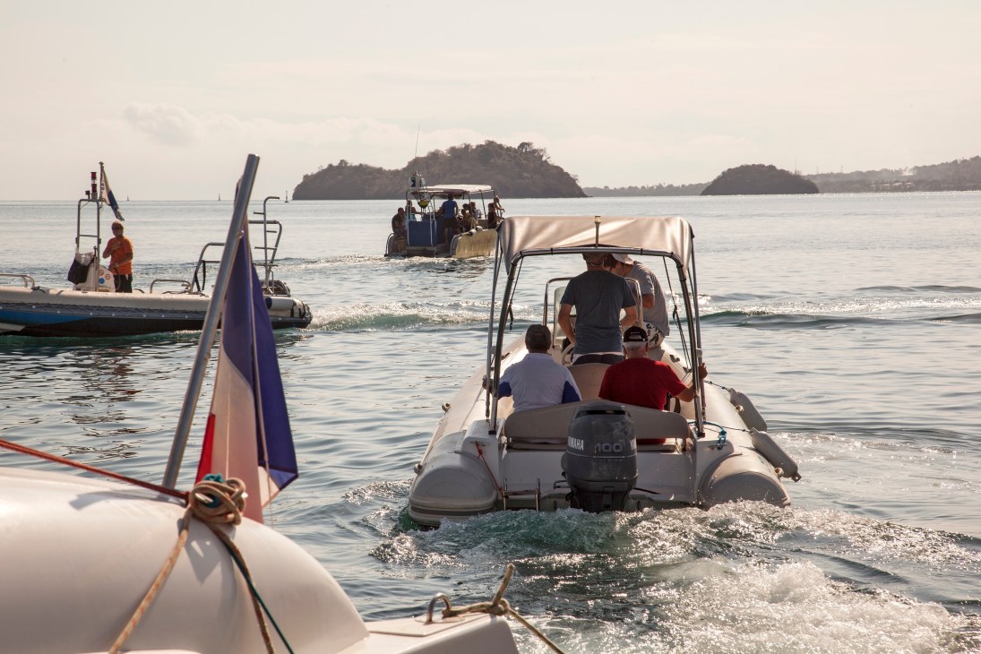 Speedboats with French flags depart from a beach in Mamoudzou, Mayotte, with forested islands visible across the water.