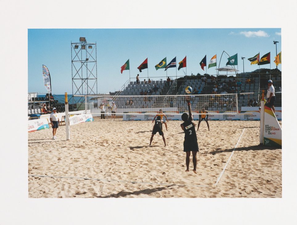 Two beach volleyball players in action during a match on sand with a net and scoreboard visible.