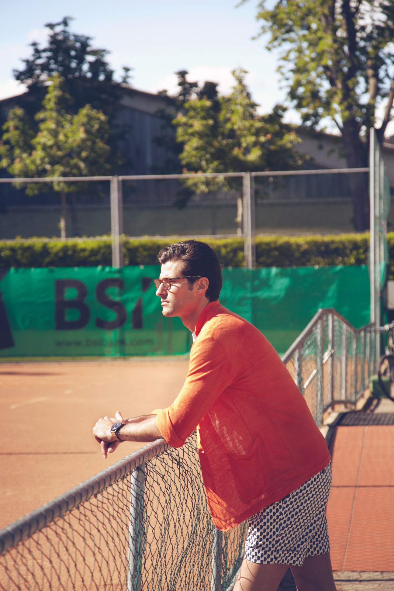 Man in red cardigan and Gucci shorts leaning against tennis net at clay court in Lugano.