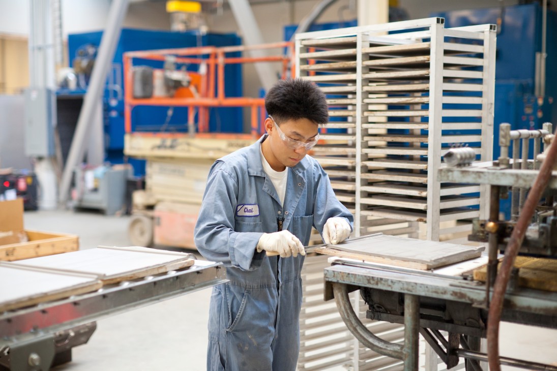 Worker in blue jumpsuit inspecting ceramic tiles in a factory with blue shelving racks and machinery in background.