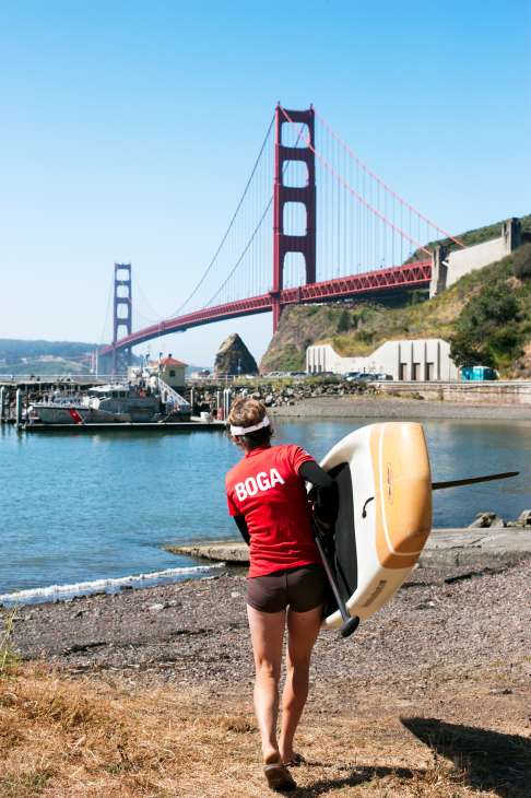 A woman carrying a paddleboard walks on a rocky beach with the Golden Gate Bridge visible in the background.