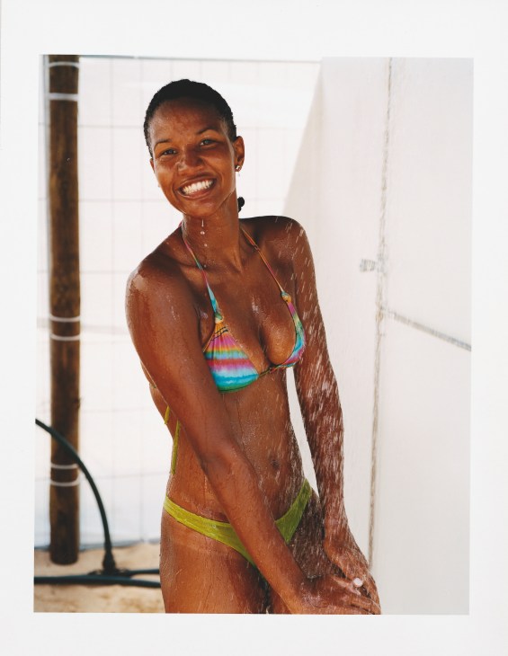 Woman in colorful striped bikini wet from water spray, smiling at camera during beach volleyball.