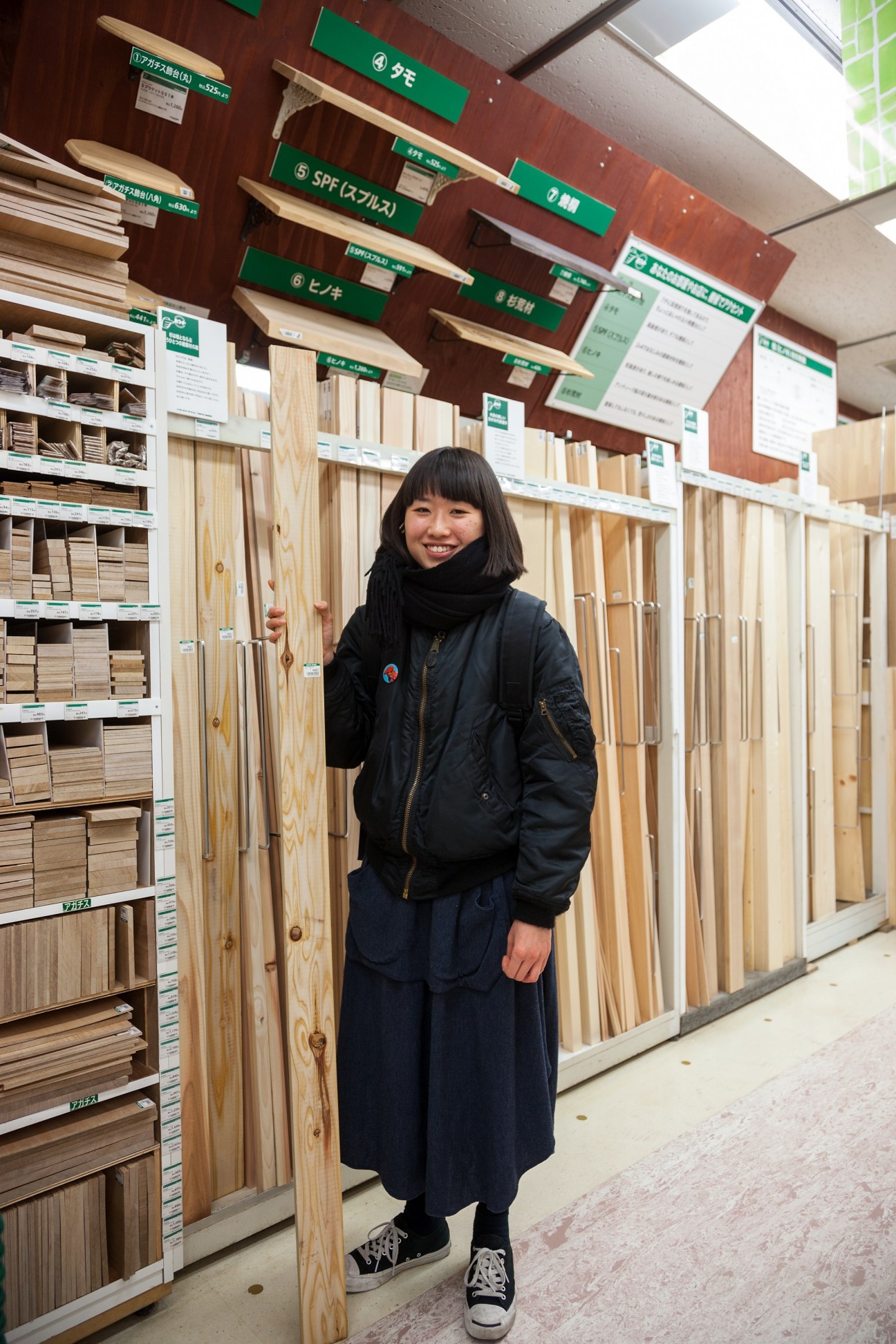 Young woman smiling in woodwork department surrounded by wood planks and material samples.