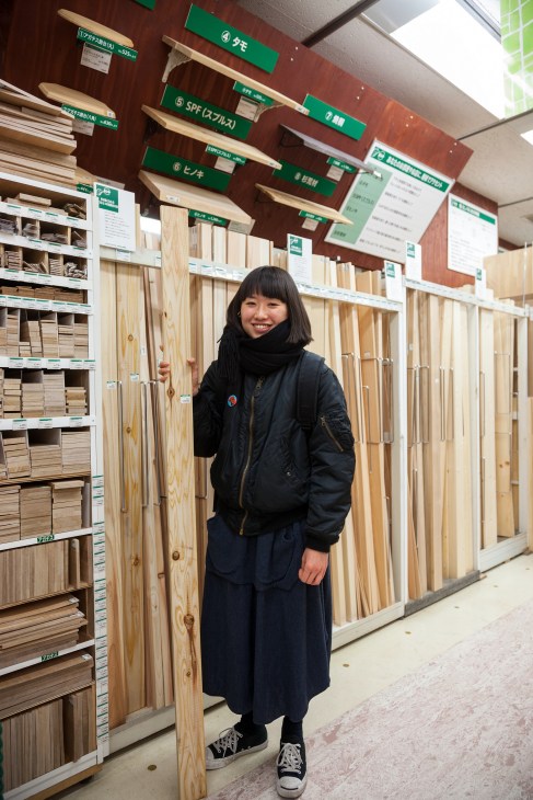 Young woman smiling in woodwork department surrounded by wood planks and material samples.
