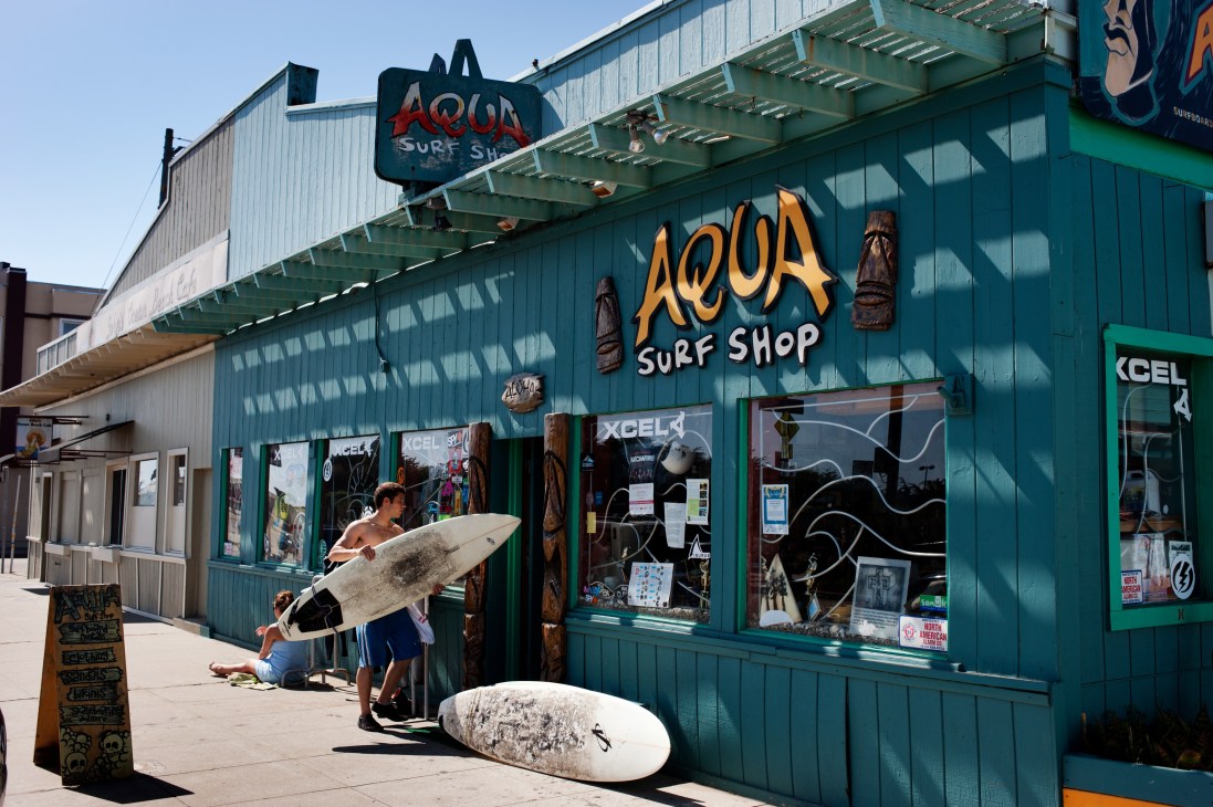 Surfer carrying board outside Aqua Surf Shop in San Francisco with teal storefront and wooden tiki decorations.