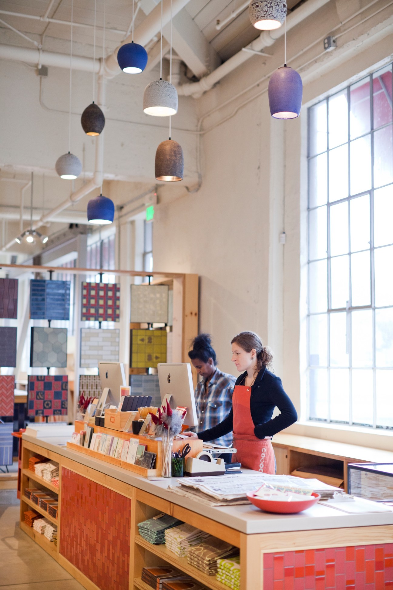 Two people work at a wooden counter in a bright industrial space with hanging ceramic pendant lights and material samples on display.