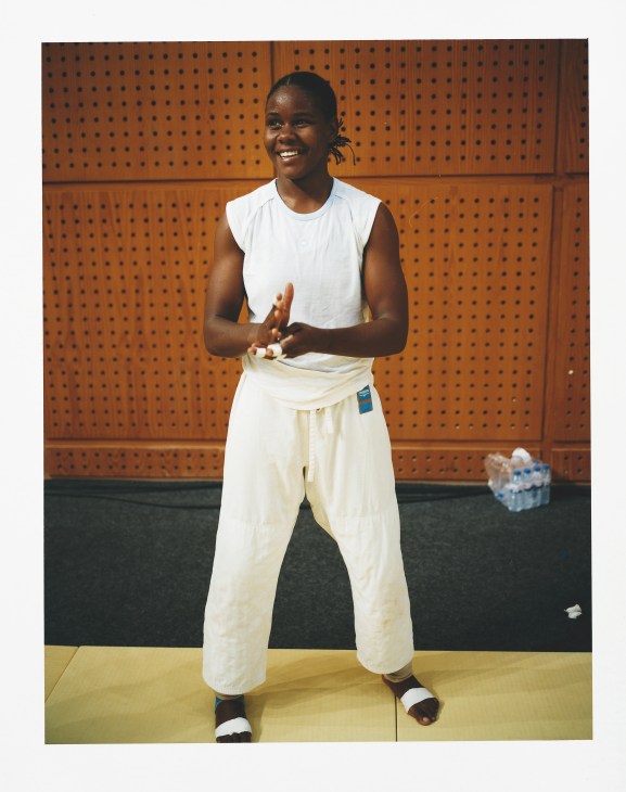 A smiling woman in a white sleeveless shirt and cream-colored pants stands against a brown perforated wall backdrop, holding rolled towels.