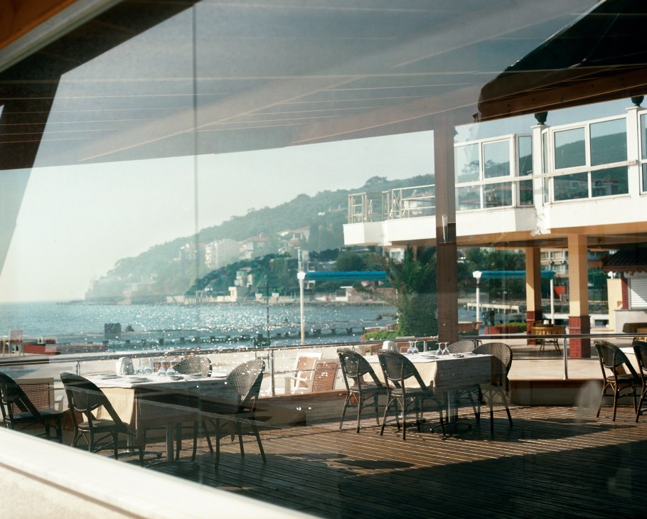 Waterfront lido with wooden terrace, dining tables, and shaded canopy overlooking turquoise sea and hillside buildings.