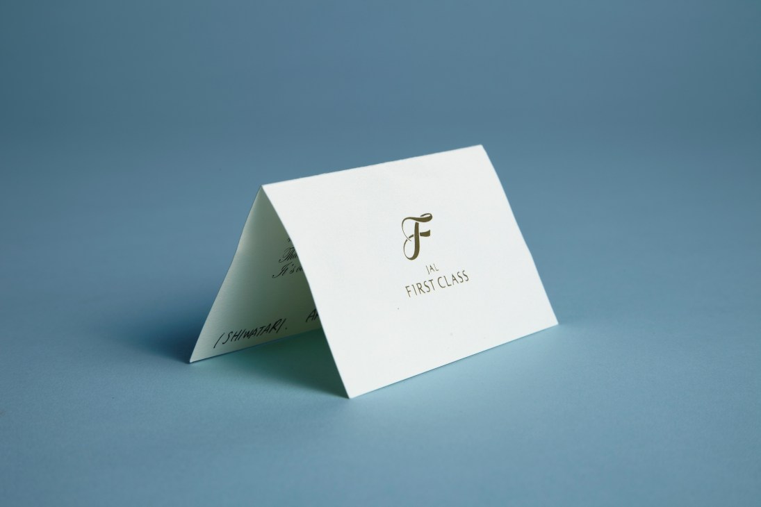 A white folded thank you card with JAL First Class logo and handwritten text on blue background.