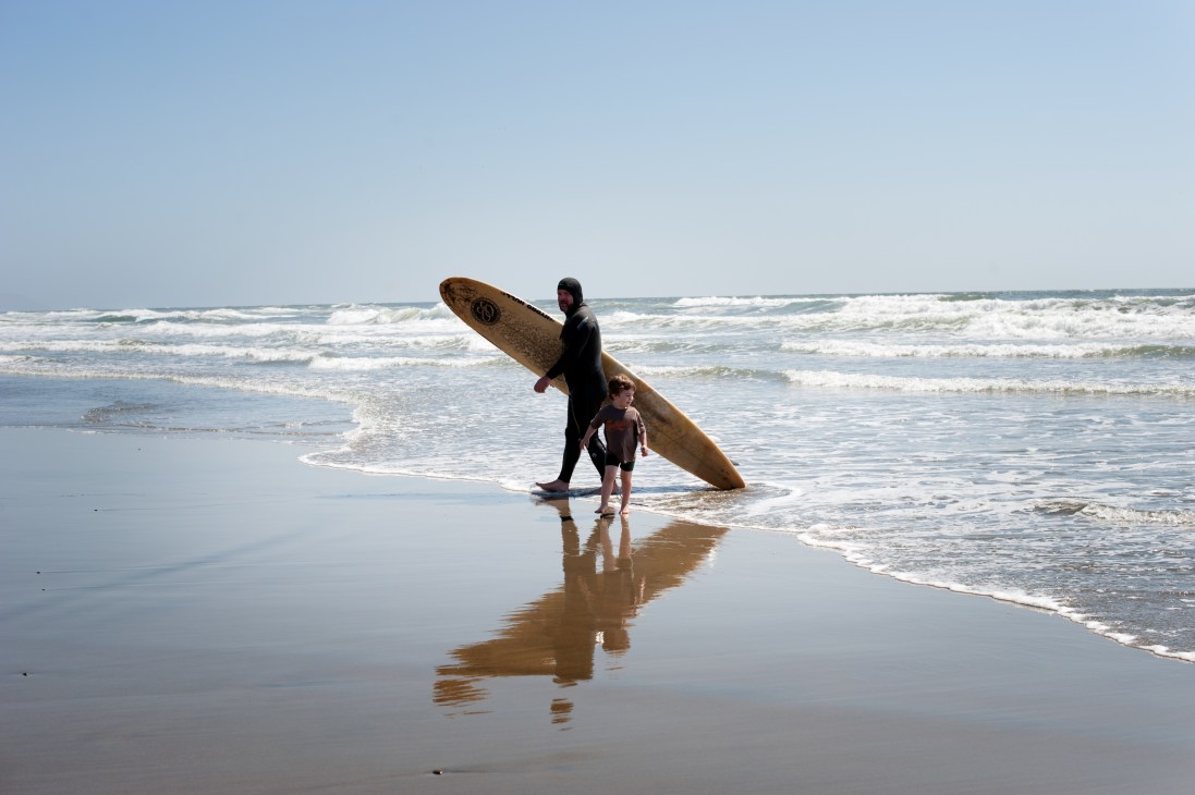 An adult surfer in a wetsuit carrying a surfboard walks with a young child on a beach with waves in the background.