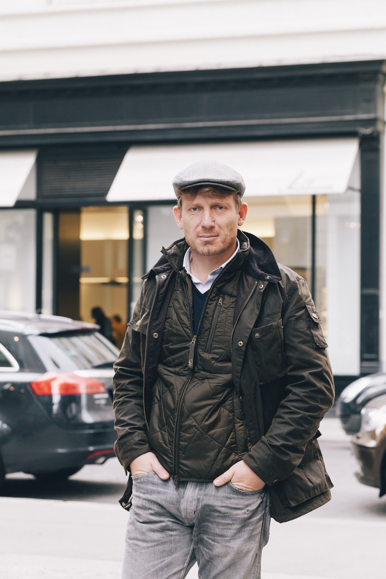 Man in black quilted jacket and flat cap standing outside storefront.