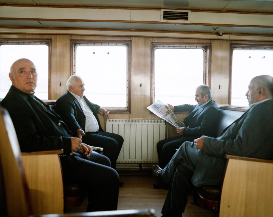 Four men sitting inside a ferry cabin reading a newspaper and conversing.