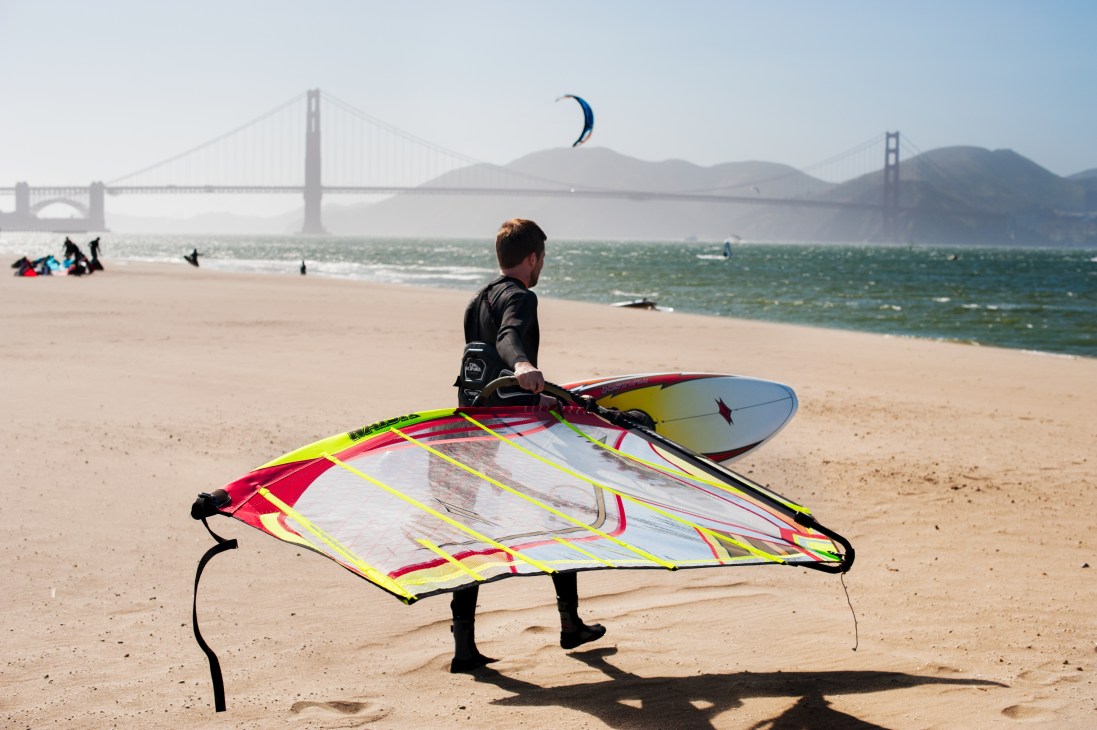 Kite surfer in wetsuit carrying colorful sail on San Francisco beach with Golden Gate Bridge visible in background.