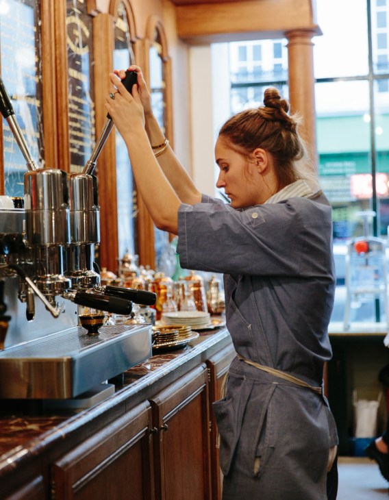 Staff member pouring coffee at Grand Café Tortoni counter with wooden interior and Parisian street view.