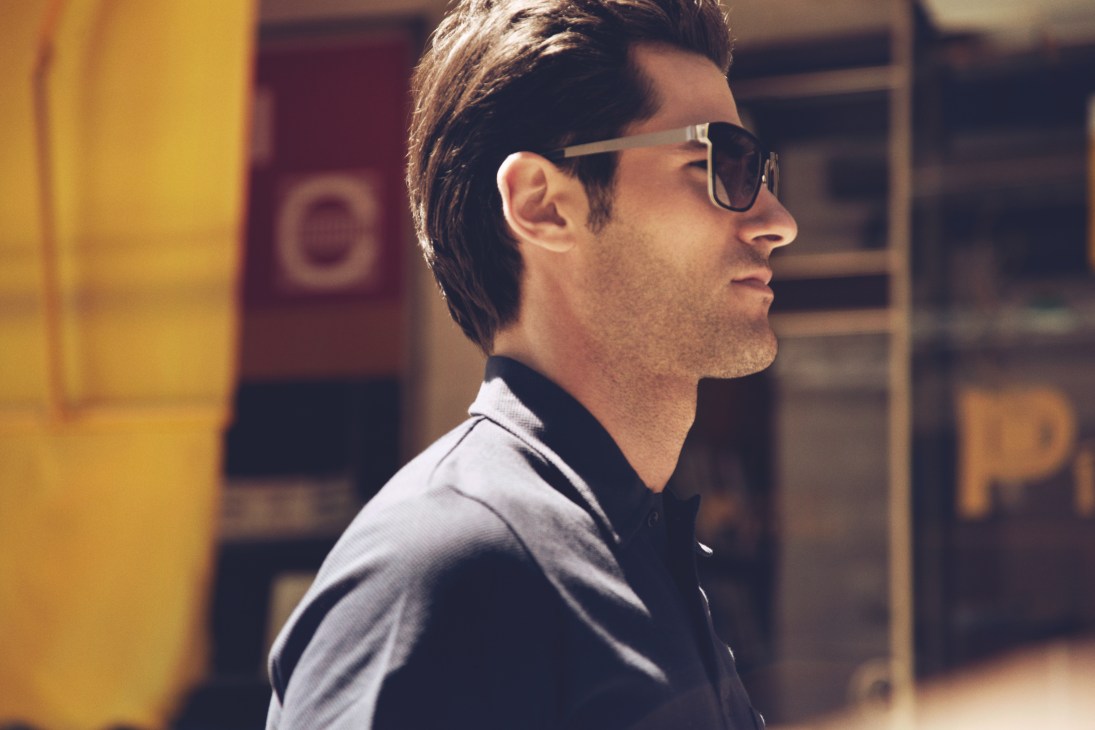 Man in profile wearing Lindberg sunglasses and Sunspel polo shirt against colorful building backdrop.