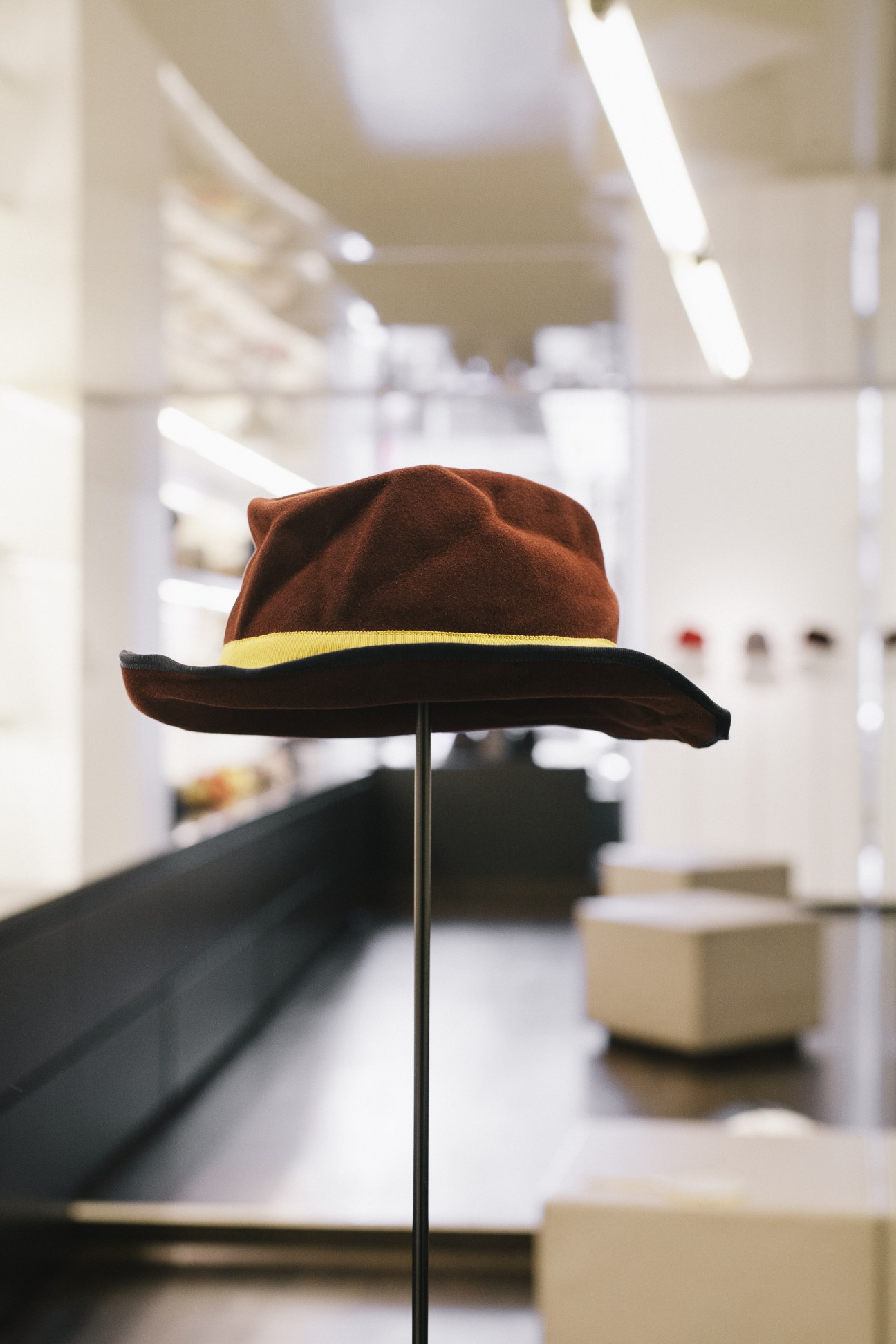 Brown felt hat with black brim and yellow band displayed on stand in Mühlbauer hat shop.