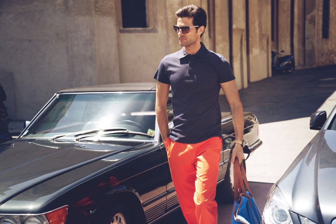 Man in sunglasses, black polo shirt and red trousers standing beside a vintage convertible car.