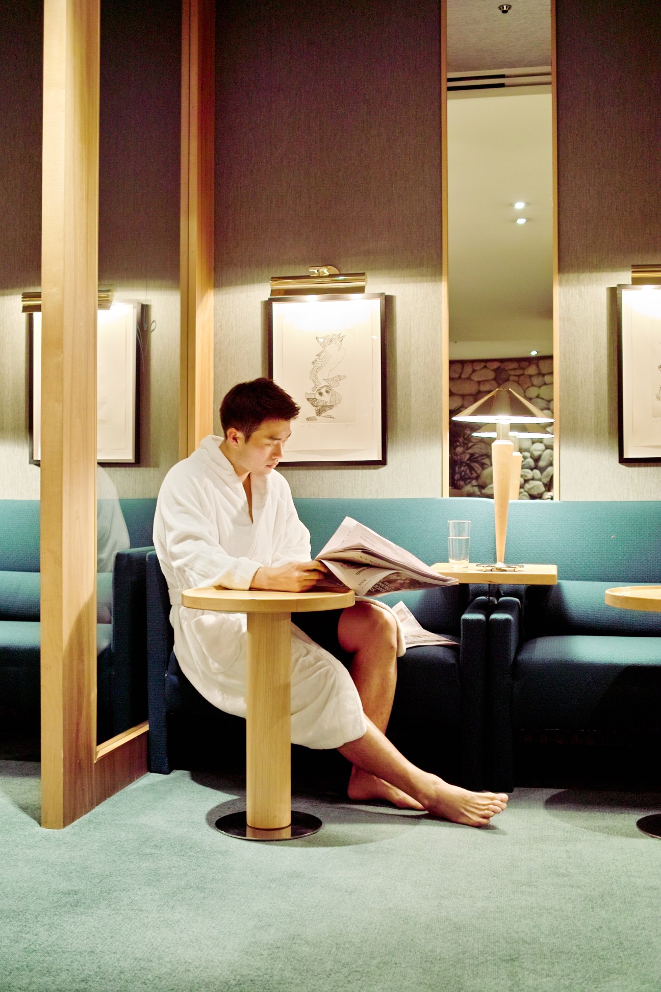 Man in white robe reading magazine in modern hotel lounge booth with turquoise seating and wood paneling.