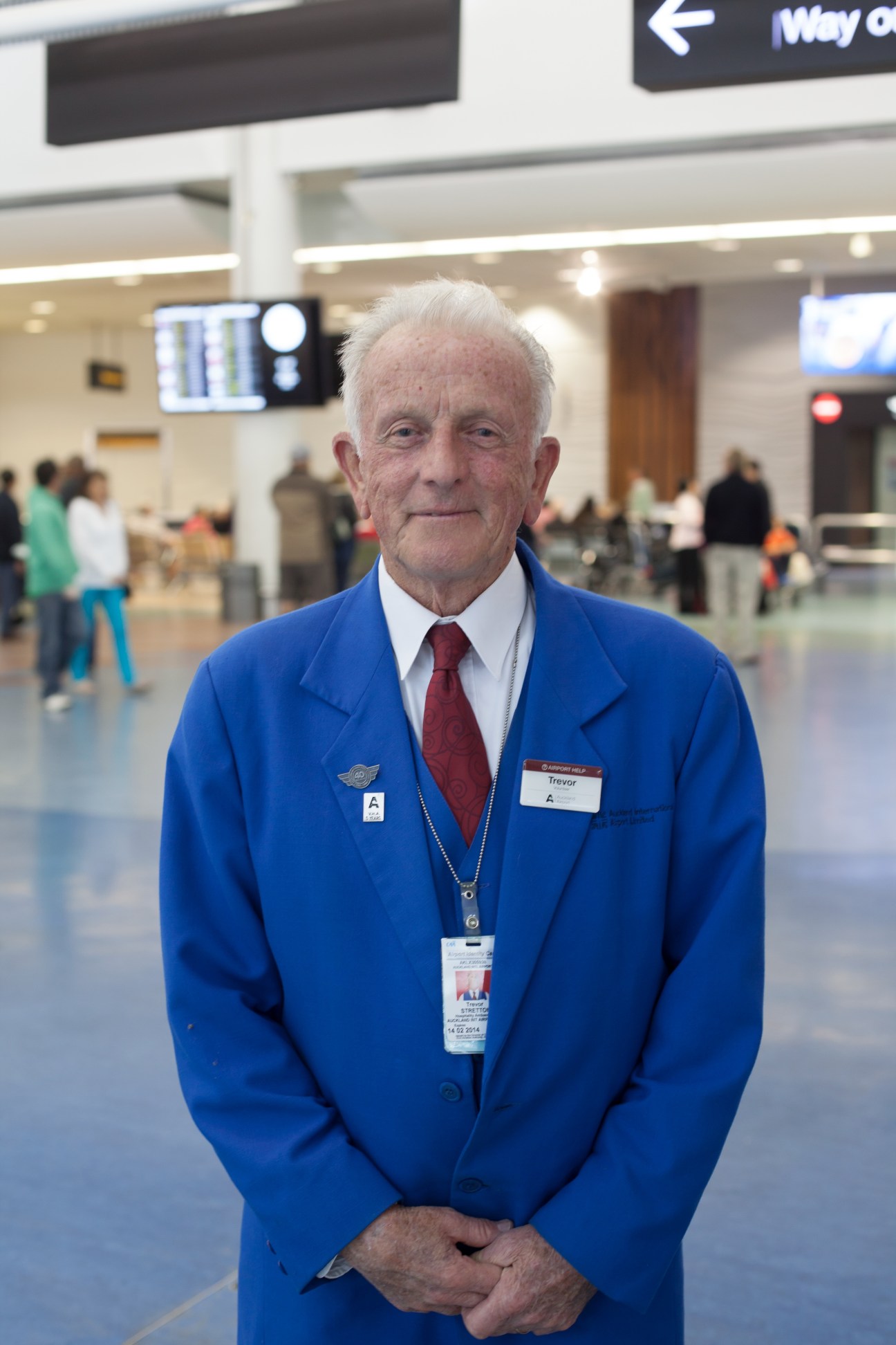 Elderly airport staff member in blue uniform and red tie at Auckland Airport terminal.