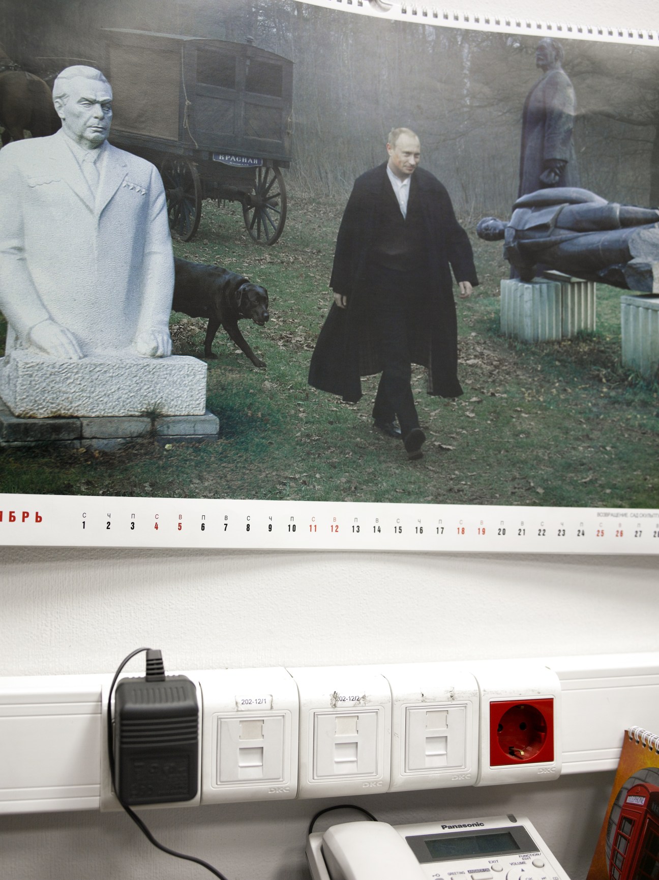 Putin calendar hanging on office wall above electrical outlets and equipment.