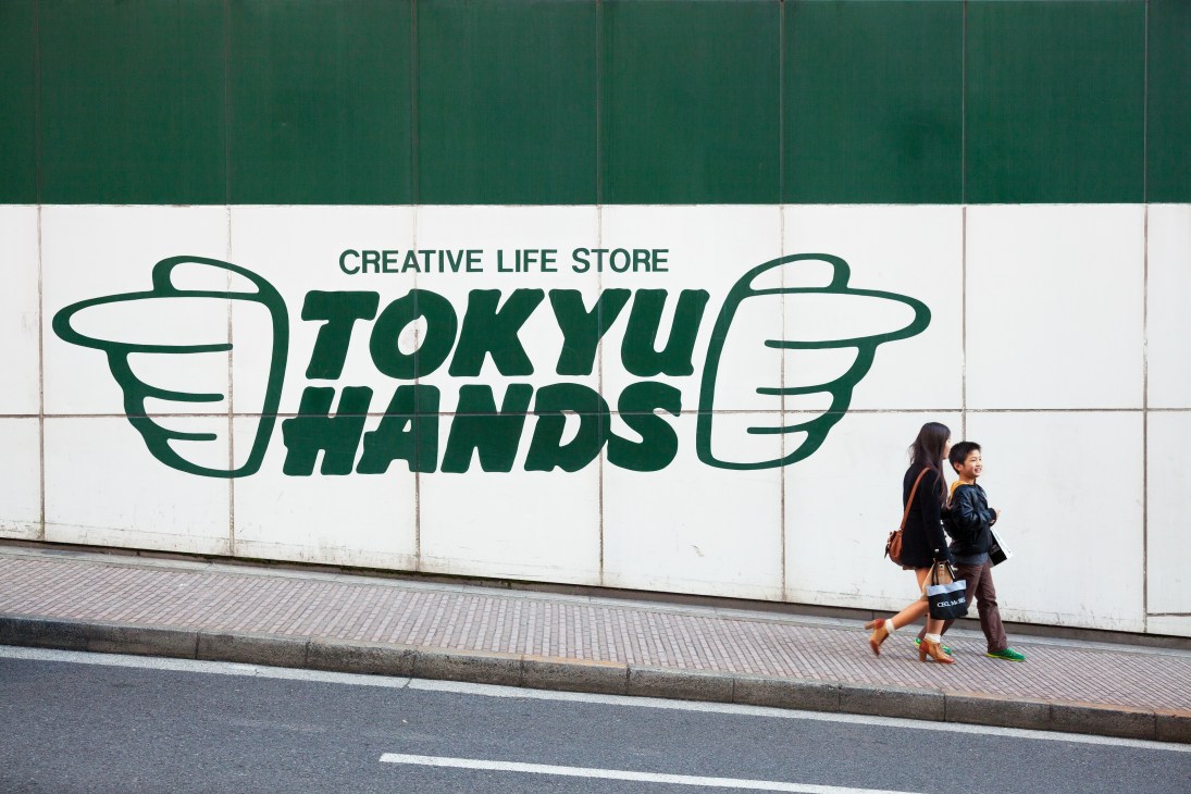 Two pedestrians walk past Tokyu Hands storefront with green and white signage displaying the store's logo of two hands and 'Creative Life Store' text.