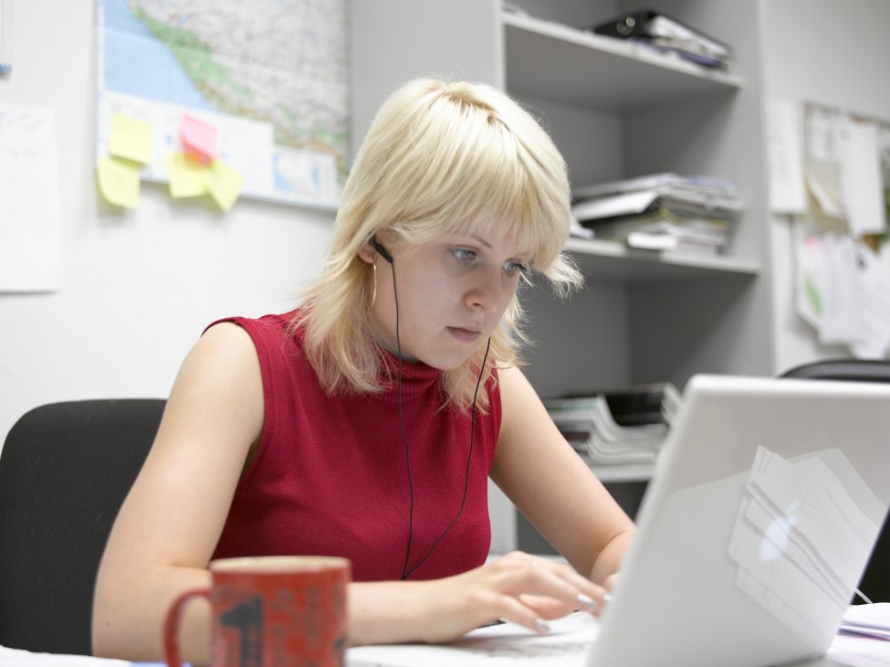 Journalist Ekaterina Bazanova wearing headphones and red sleeveless top, working at a laptop in an office.